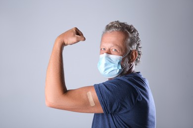 Photo of Senior man in protective mask showing arm with bandage after vaccination on grey background
