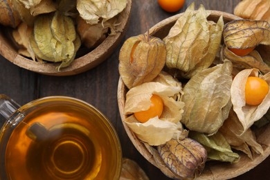 Ripe physalis fruits with calyxes in bowls and cup of tea on wooden table, flat lay