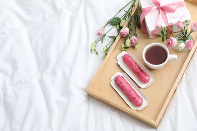 Photo of Tasty breakfast served in bed. Delicious eclairs, tea, gift box, flowers and card with phrase I Love You on tray, above view. Space for text