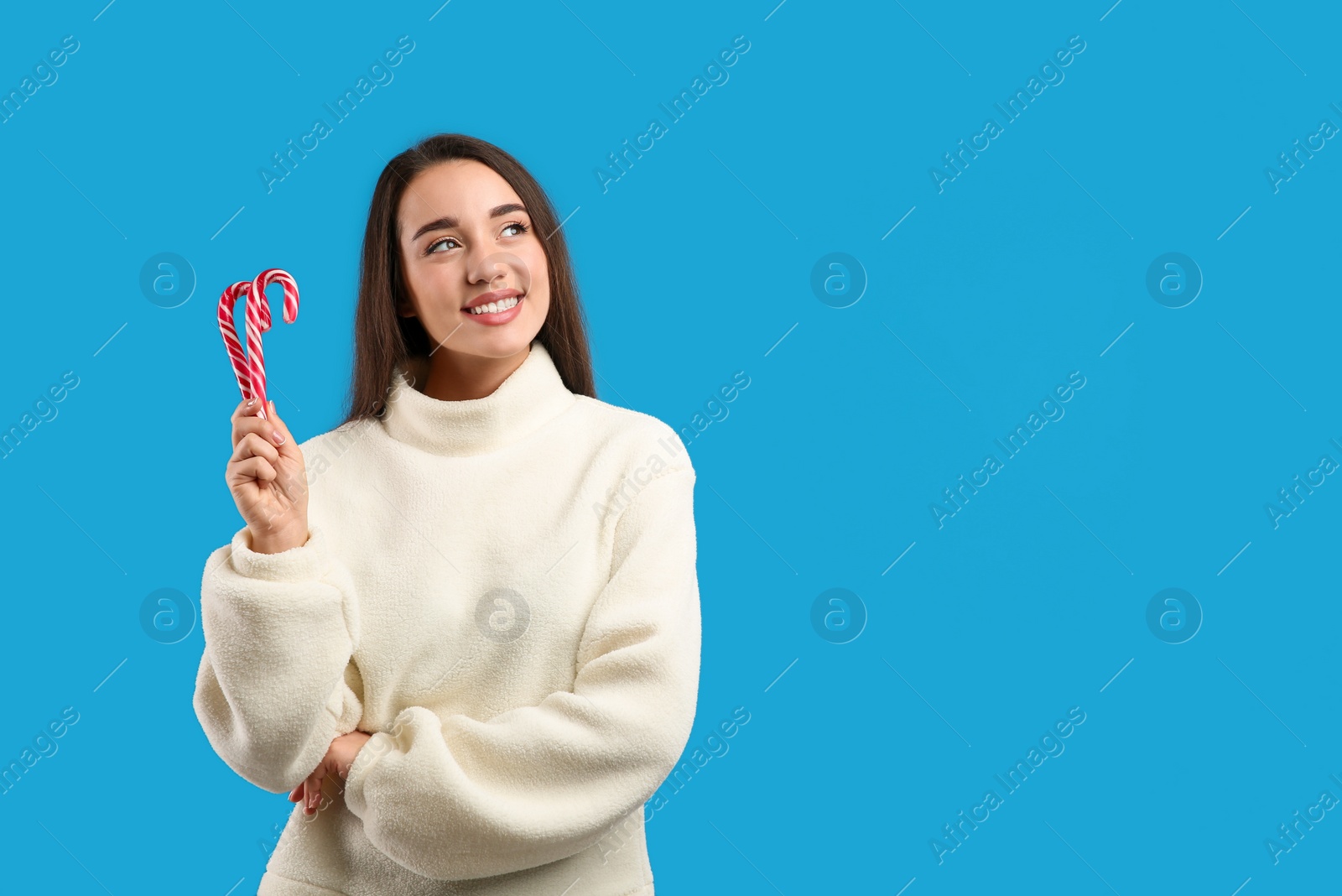 Photo of Young woman in beige sweater holding candy canes on blue background, space for text. Celebrating Christmas