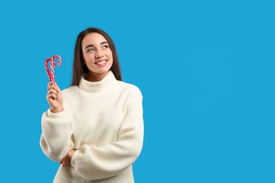 Photo of Young woman in beige sweater holding candy canes on blue background, space for text. Celebrating Christmas
