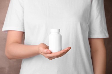 Photo of Woman holding blank white bottle with vitamin pills against light brown background, closeup