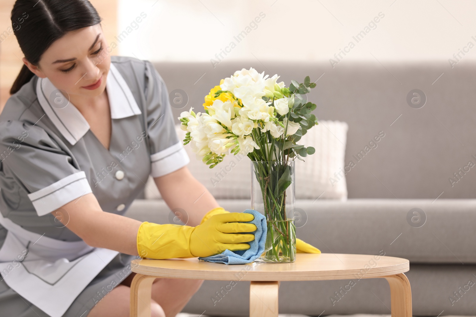 Photo of Young chambermaid wiping dust from wooden table in hotel room