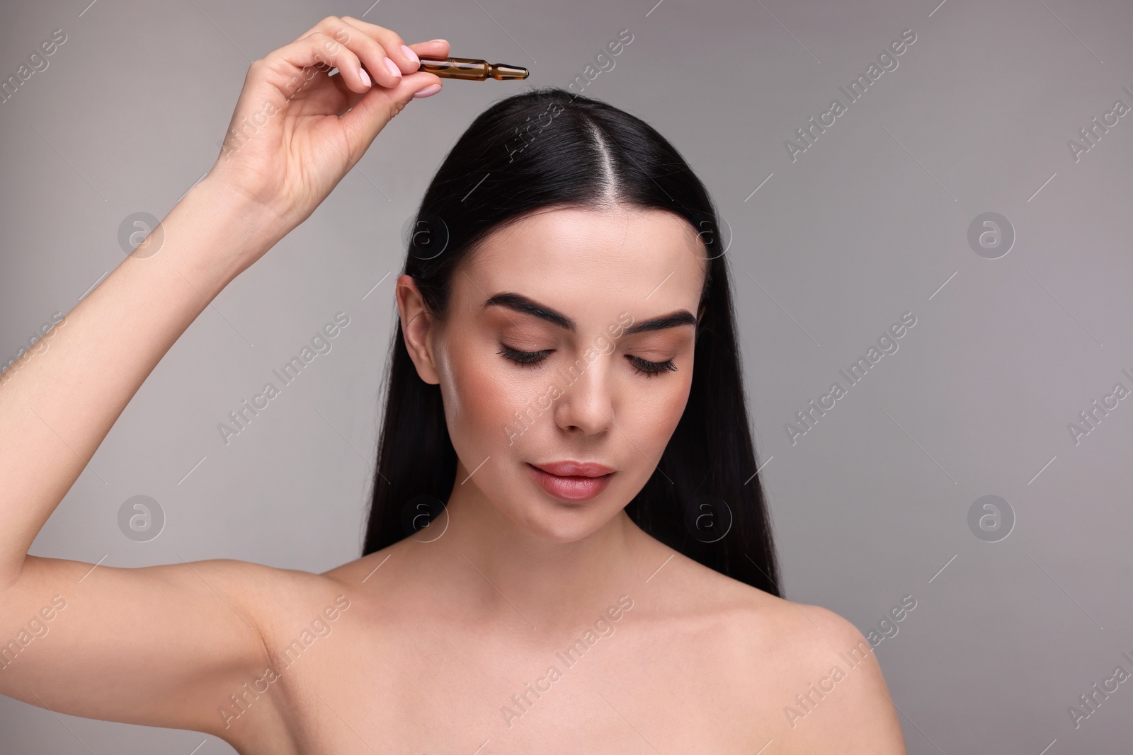 Photo of Beautiful young woman using ampoule for hair treatment on grey background