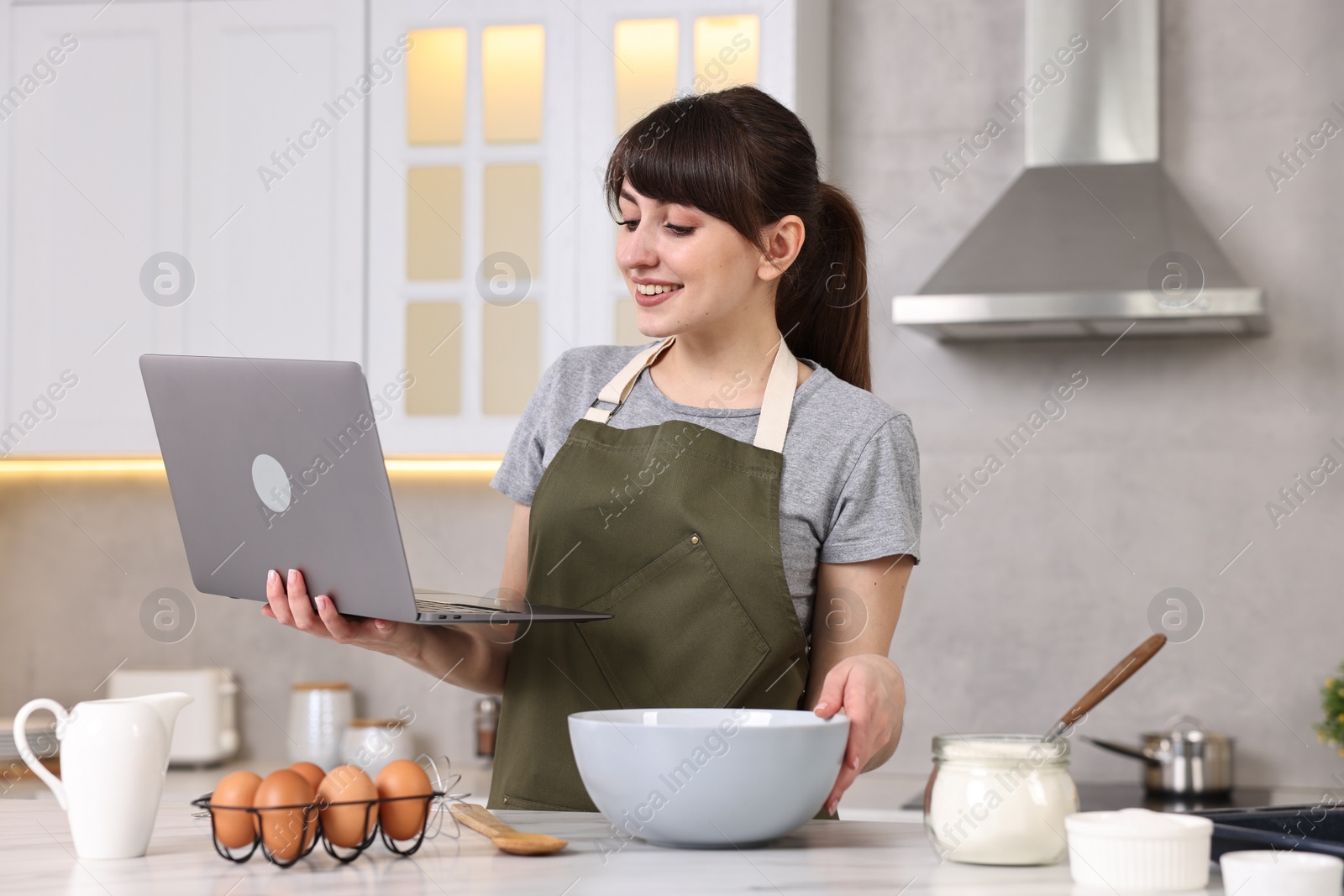 Photo of Happy young housewife using laptop while cooking at white marble table in kitchen