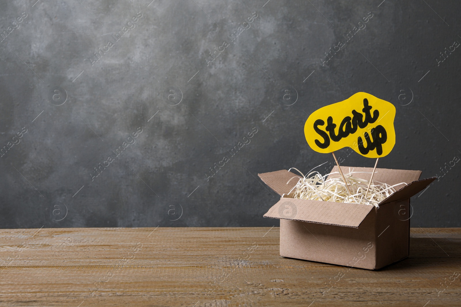 Photo of Open cardboard box and sign with words Start Up on wooden table against grey background, space for text