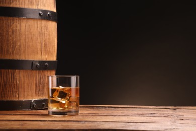 Photo of Whiskey with ice cubes in glass and barrel on wooden table against black background, space for text