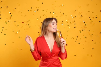 Happy young woman with glass of champagne and confetti on yellow background