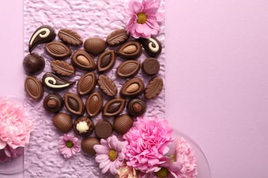 Heart made with delicious chocolate candies and beautiful flowers on pink background, flat lay. Space for text