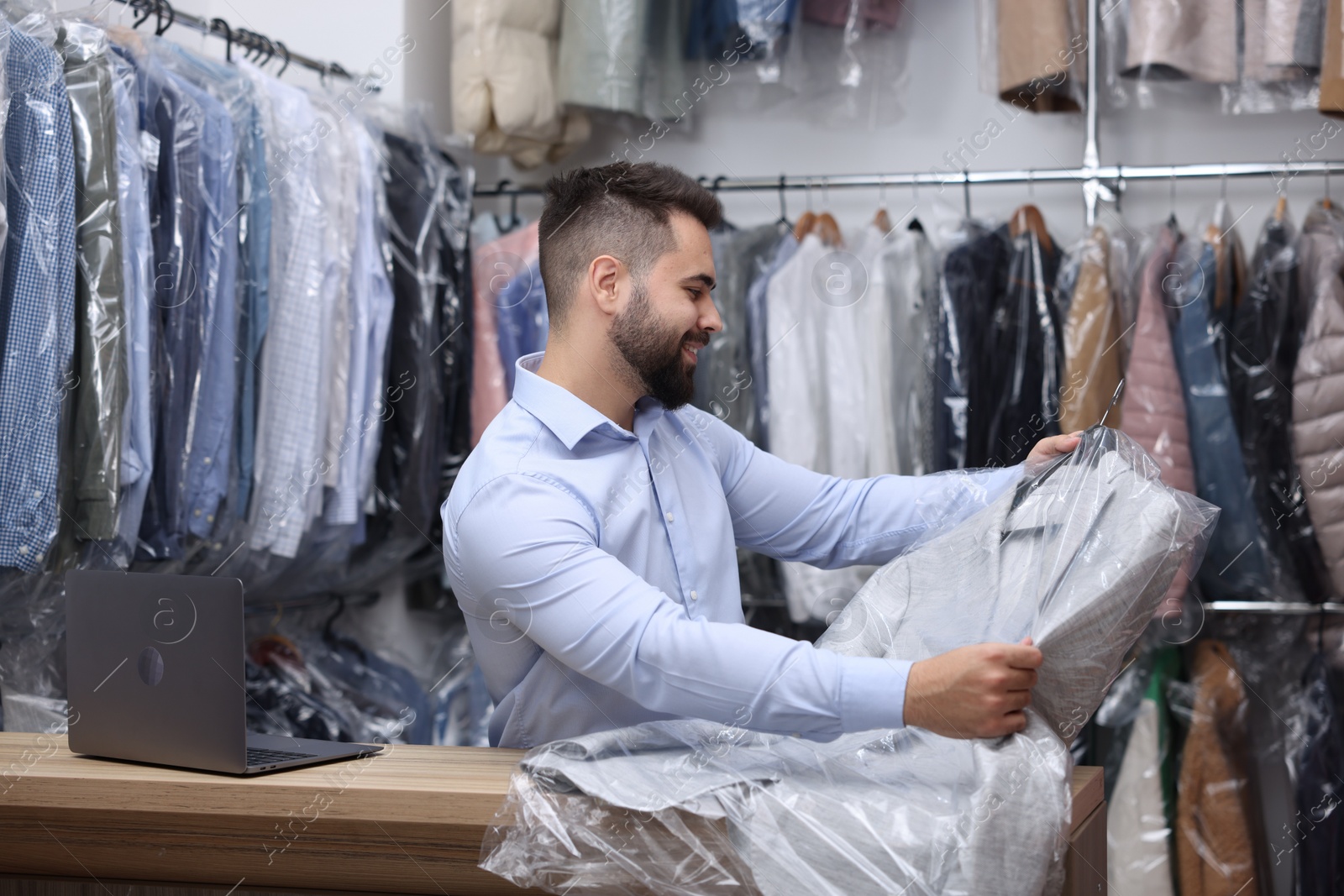 Photo of Dry-cleaning service. Happy worker holding hanger with jacket in plastic bag at counter indoors