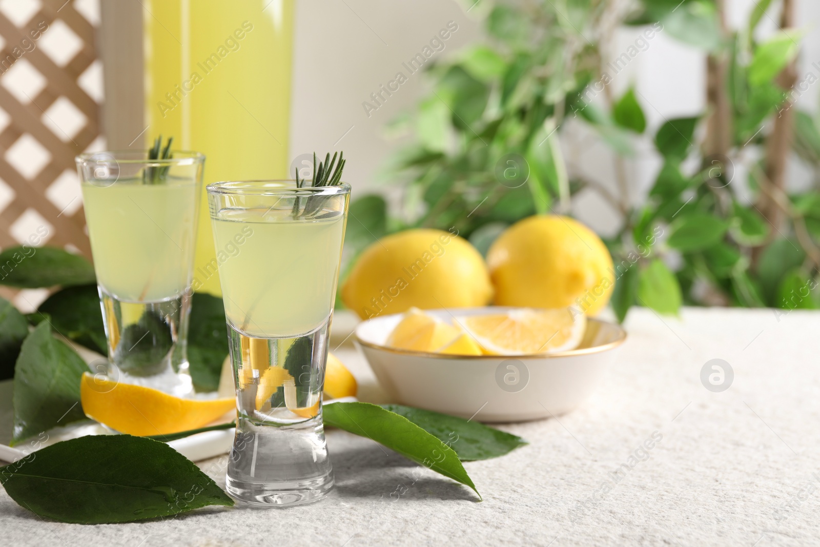 Photo of Tasty limoncello liqueur, lemons and green leaves on white textured table, closeup. Space for text