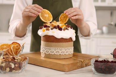 Photo of Woman decorating traditional Easter cake with dried orange slices at white marble table in kitchen, closeup