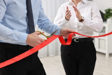 Photo of Man cutting red ribbon with scissors indoors, closeup