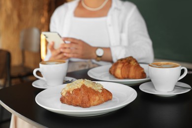 Photo of Woman sitting near tasty pastry and cups of aromatic coffee at black table in cafeteria, focus on croissants