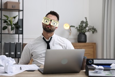 Photo of Man with fake eyes painted on sticky notes at workplace in office. Overwhelmed by work