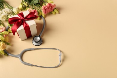 Photo of Stethoscope, gift box and flowers on beige background, space for text. Happy Doctor's Day
