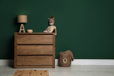 Modern wooden chest of drawers with lamp and toy near green wall indoors. Space for text