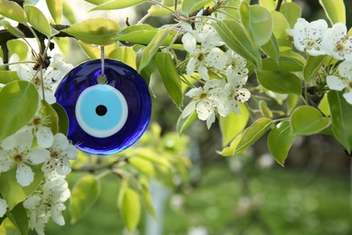 Photo of Evil eye amulet hanging on blossoming spring tree outdoors, closeup. Space for text