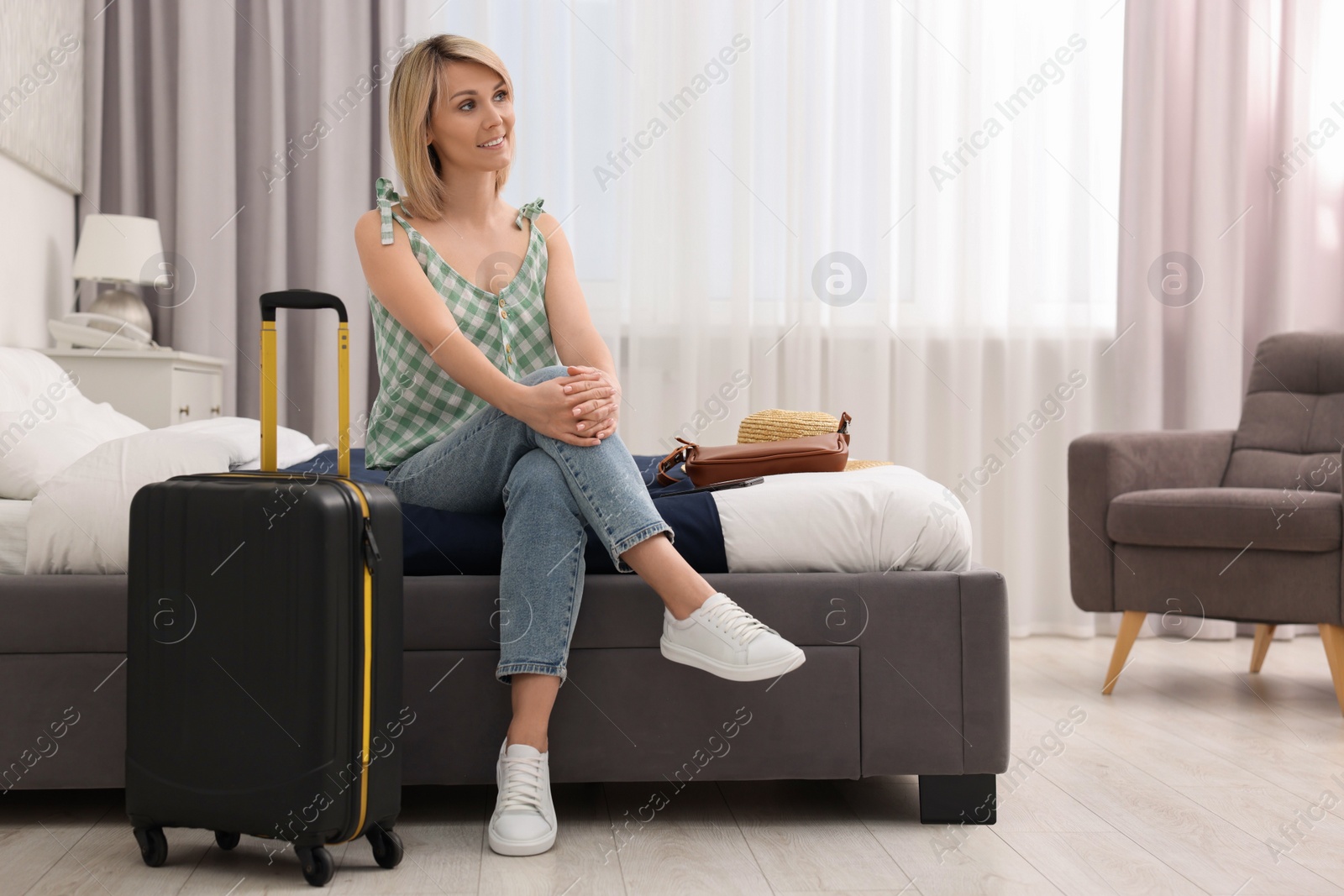 Photo of Smiling guest with suitcase relaxing on bed in stylish hotel room
