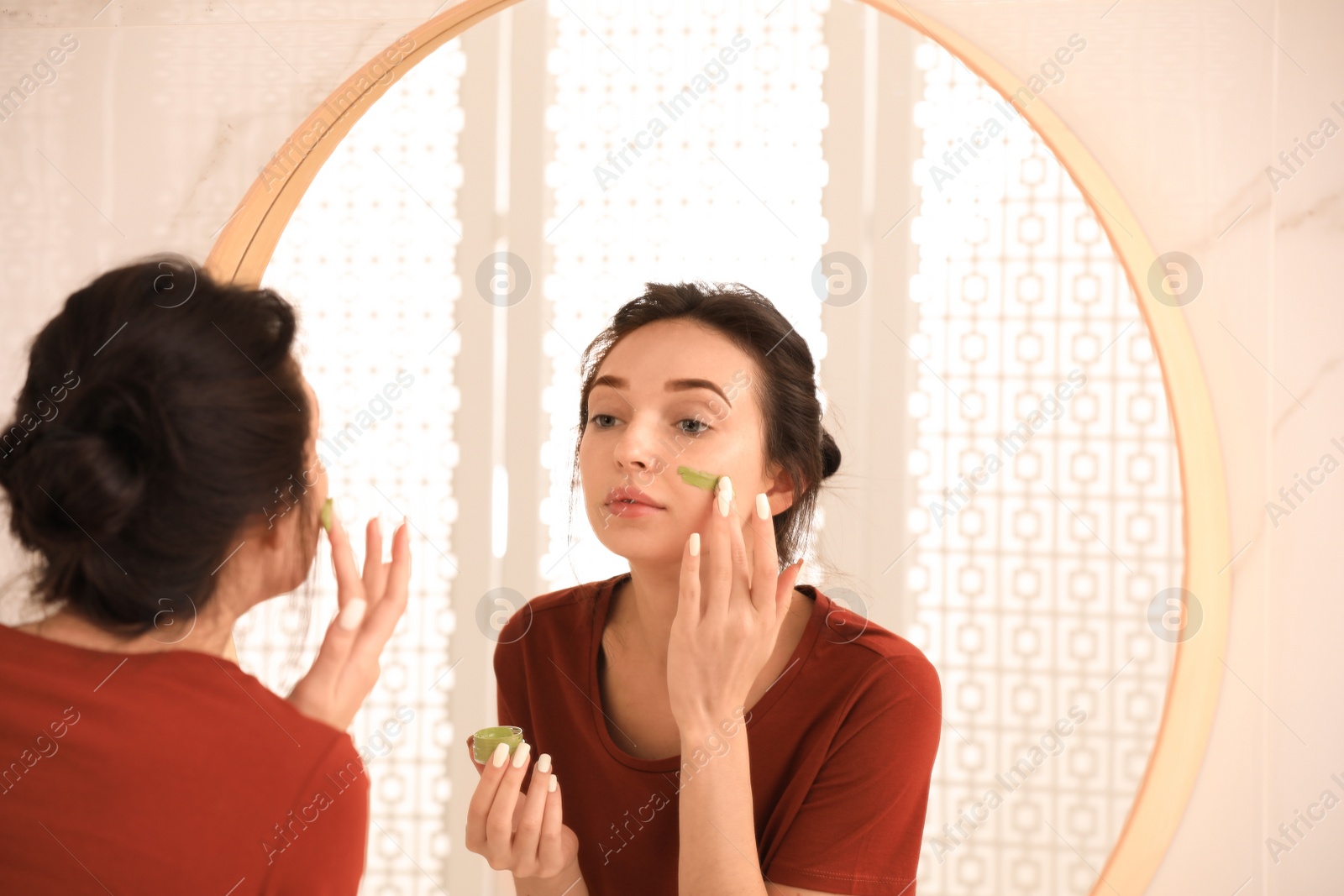 Photo of Young woman applying mask on her face near mirror in bathroom