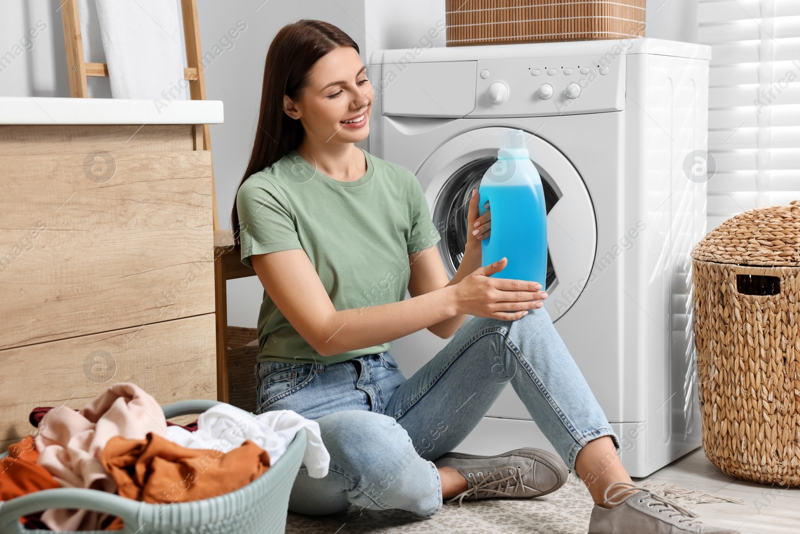 Photo of Woman sitting on floor near washing machine and holding fabric softener in bathroom