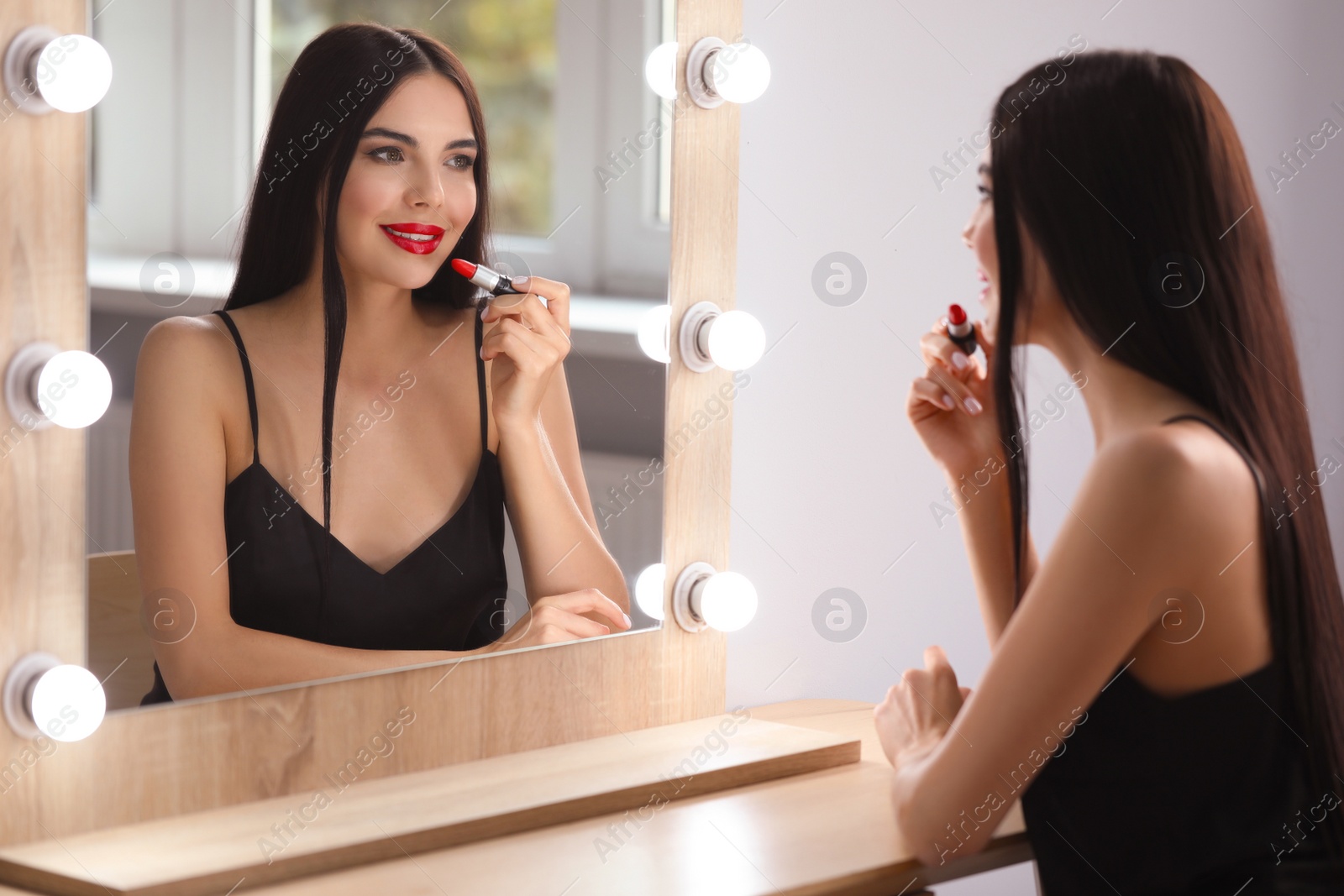 Photo of Young woman applying beautiful red lipstick in front of mirror indoors