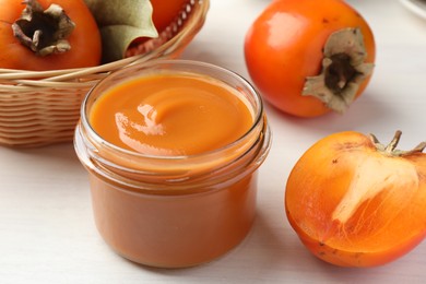 Delicious persimmon jam in glass jar and fresh fruits on white wooden table, closeup