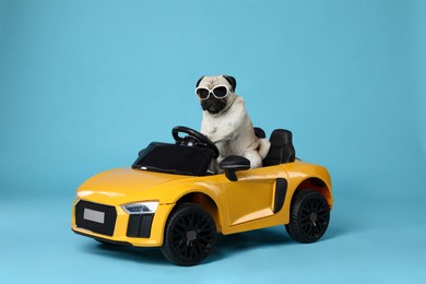 Photo of Funny pug dog with sunglasses in toy car on light blue background