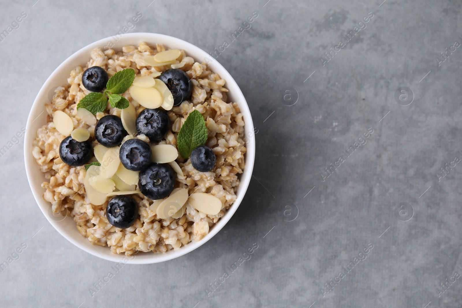 Photo of Tasty oatmeal with blueberries, mint and almond petals in bowl on grey table, top view. Space for text