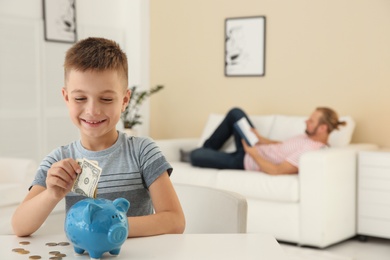 Boy putting money into piggy bank on table indoors. Space for text