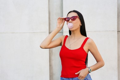 Beautiful woman in stylish sunglasses blowing gum near wall outdoors, space for text