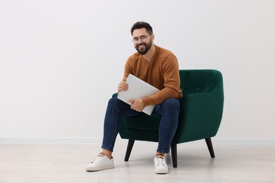 Photo of Handsome man with laptop sitting in armchair near white wall indoors, space for text