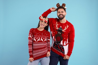 Photo of Happy young couple in Christmas sweaters, reindeer headband and funny glasses on light blue background
