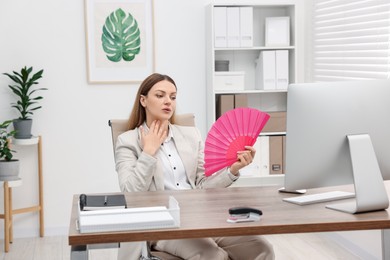 Photo of Businesswoman waving pink hand fan to cool herself at table in office