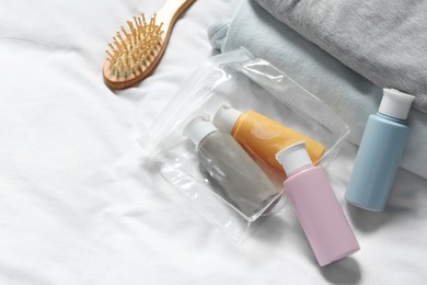 Photo of Cosmetic travel kit. Plastic bag with small containers of personal care products, brush and stack of clothes on bed