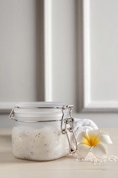 Photo of Body scrub in glass jar, towel and plumeria flowers on white wooden table, space for text