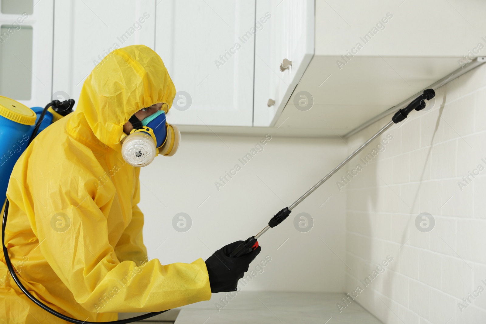 Photo of Pest control worker in protective suit spraying insecticide on furniture indoors