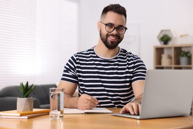 Young man writing in notebook while working on laptop at wooden table indoors