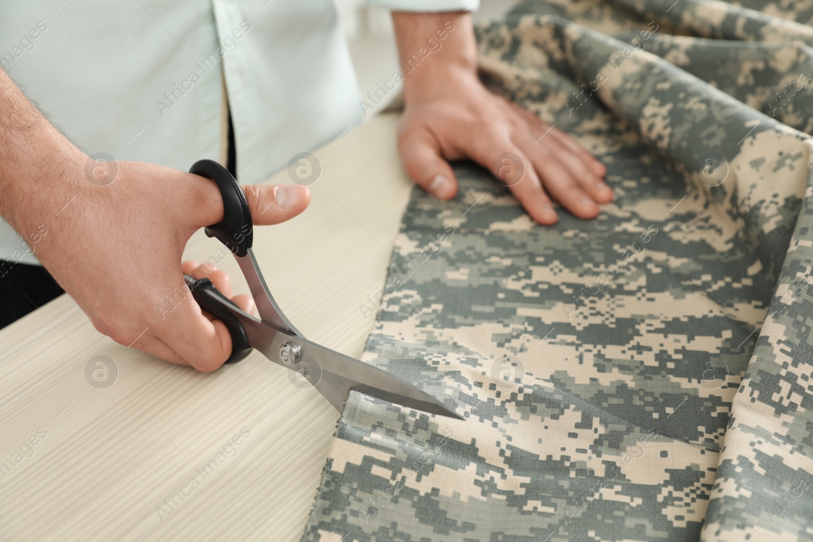 Photo of Professional tailor cutting camouflage fabric with scissors in workshop, closeup