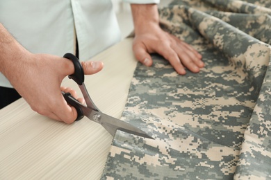 Photo of Professional tailor cutting camouflage fabric with scissors in workshop, closeup