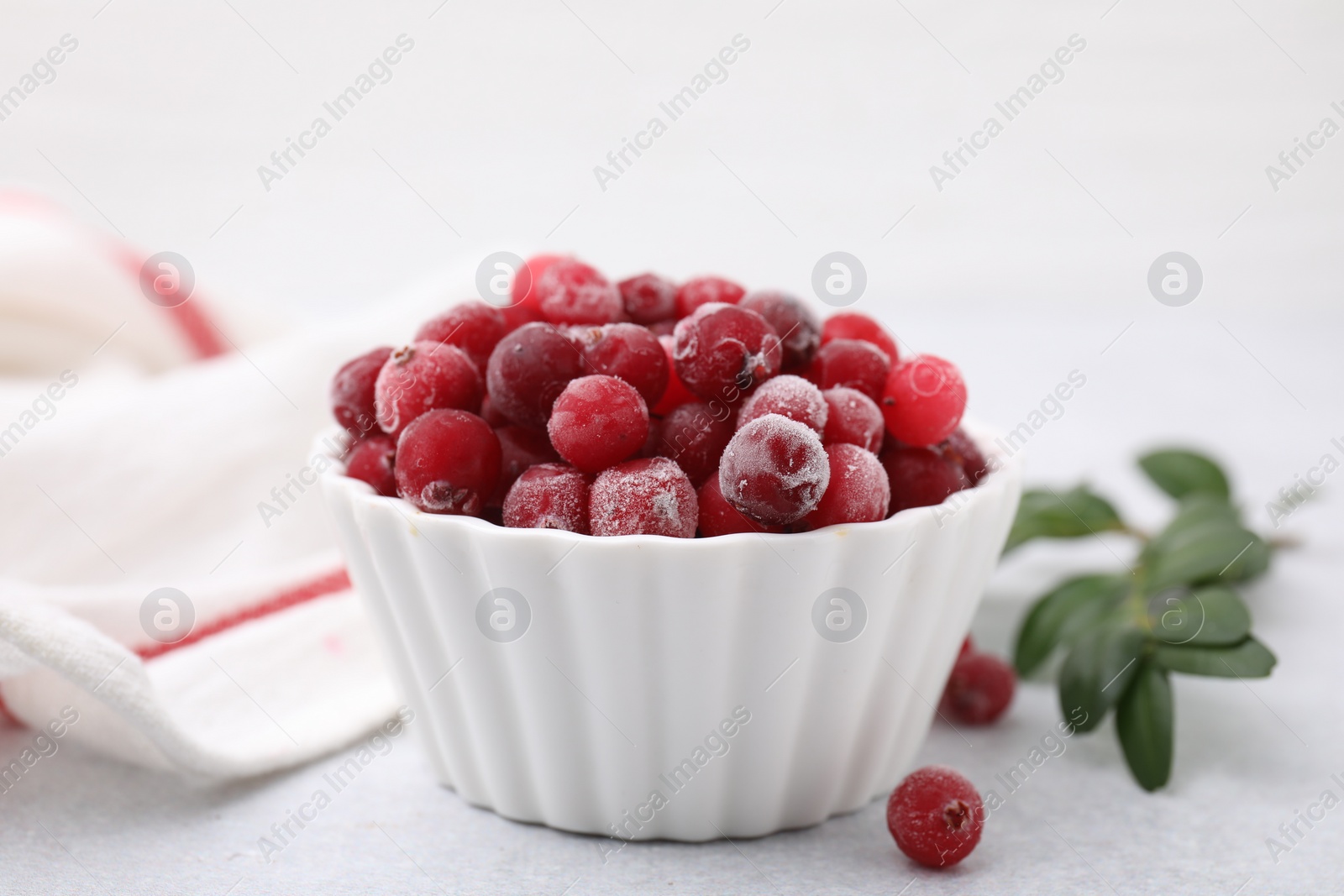 Photo of Frozen red cranberries in bowl and green leaves on light table, closeup