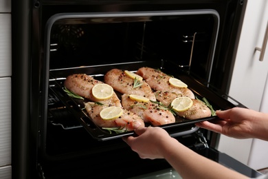 Photo of Woman putting chicken breasts with lemon and rosemary into oven, closeup