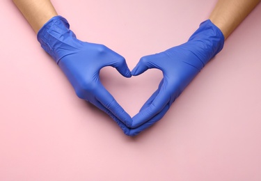 Person in medical gloves showing heart gesture against pink background, closeup of hands