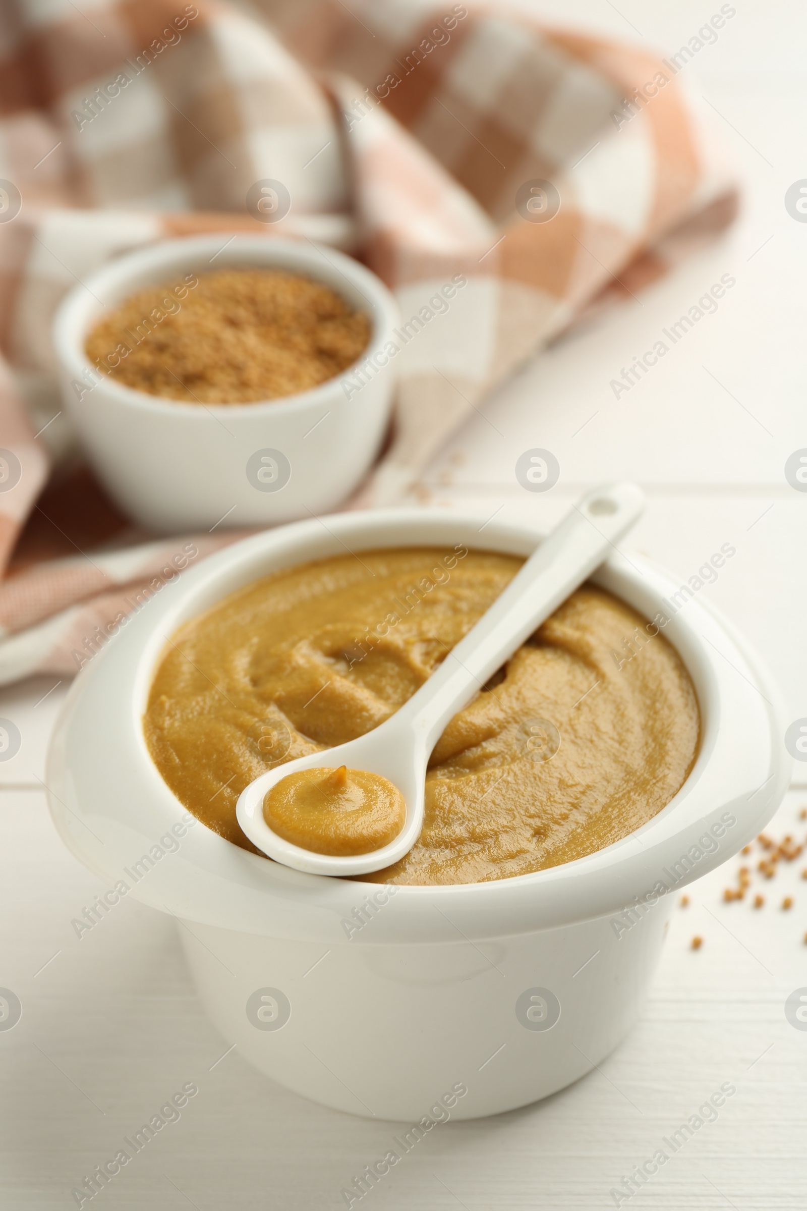 Photo of Bowl and spoon with tasty mustard sauce on white wooden table, closeup