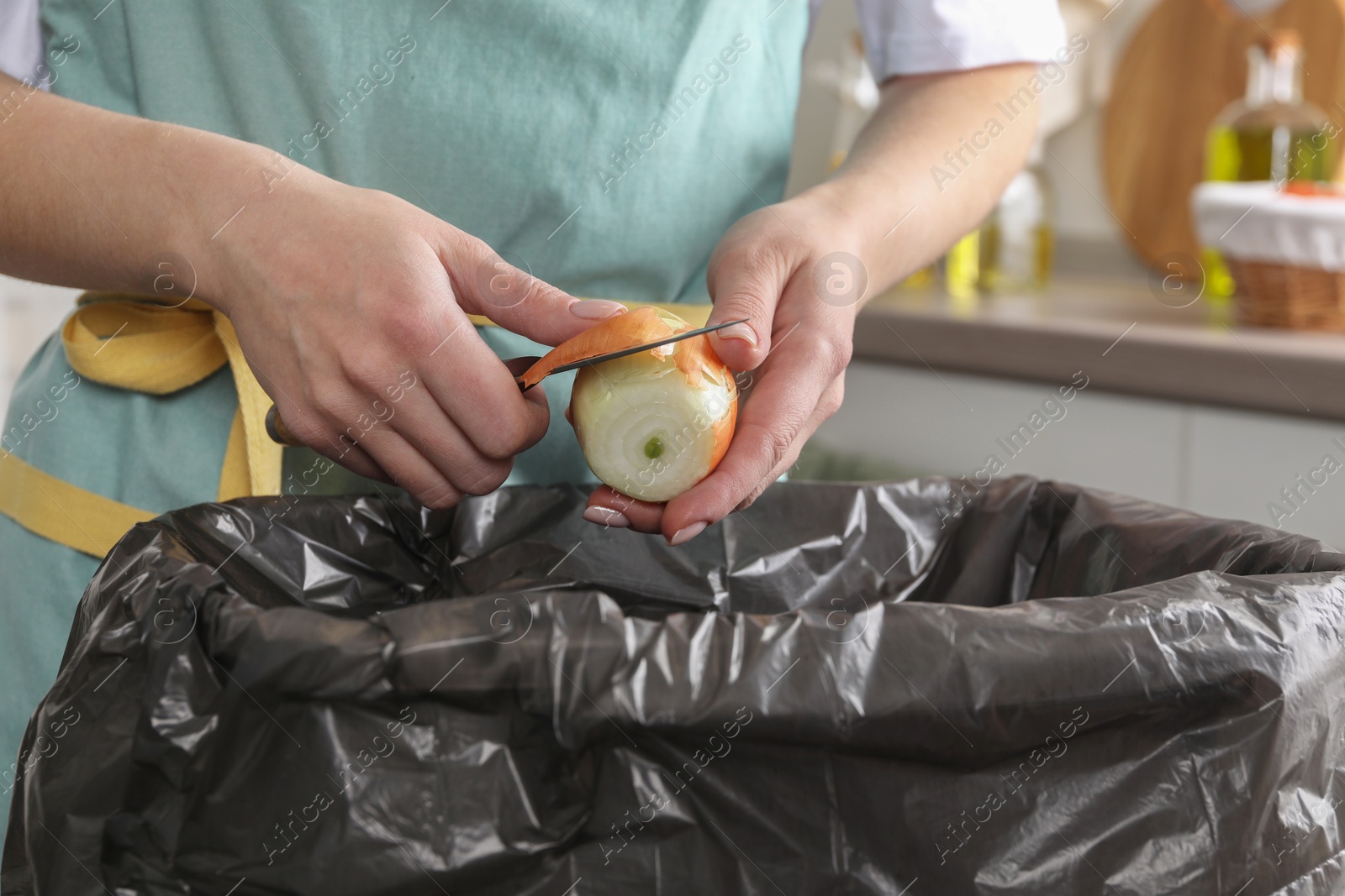 Photo of Woman peeling fresh onion with knife above garbage bin indoors, closeup