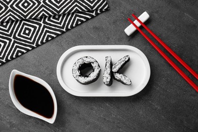 Photo of Word Ok made of sushi rolls, chopsticks and soy sauce on dark grey table, flat lay