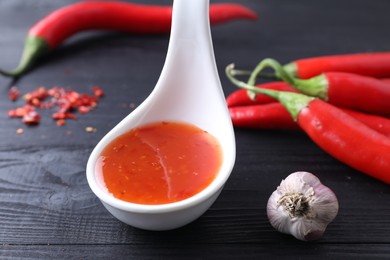 Photo of Spicy chili sauce in spoon, peppers and garlic on black wooden table, closeup