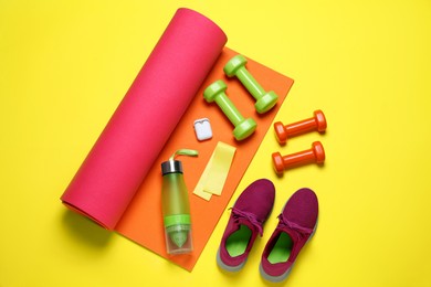 Photo of Exercise mat, dumbbells, bottle of water, wireless earphones, fitness elastic band and shoes on yellow background, flat lay