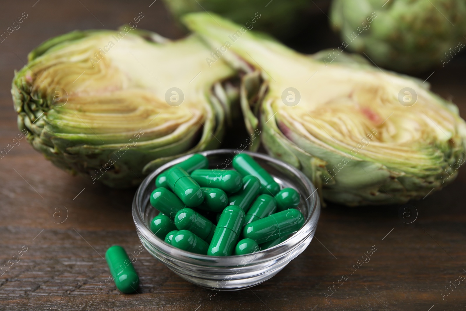 Photo of Bowl with pills and fresh artichokes on wooden table, closeup