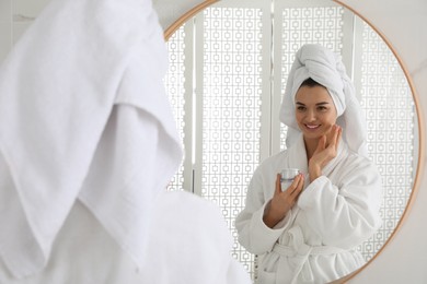 Photo of Beautiful young woman with hair wrapped in towel applying cream near mirror indoors
